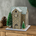 Model of a stone church with snow on the roof, surrounded by small trees and decorative elements on a wooden surface.