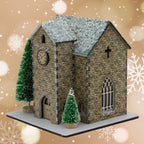 Model of a stone church with snow on the roof, surrounded by Christmas trees on a decorative background.