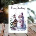 Christmas card with a snowman, birdhouse, and bird on a wooden surface with a blurred Christmas tree background.