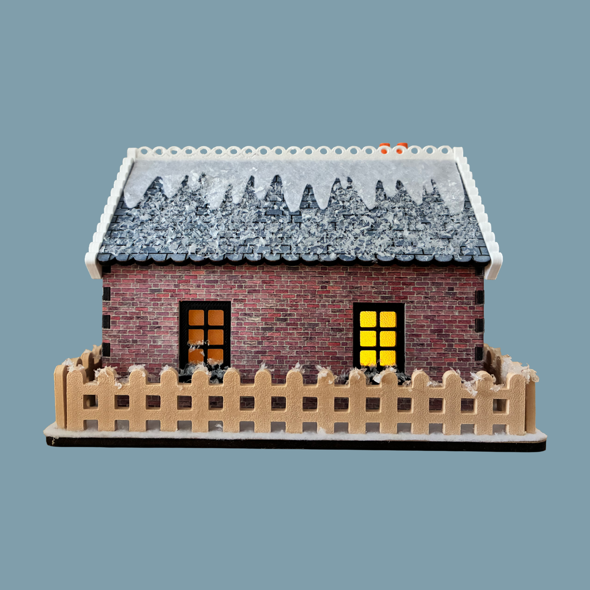A miniature brick house model with a snow-covered gray roof, two glowing yellow windows, and a beige picket fence around it, shown on a plain blue-gray background.