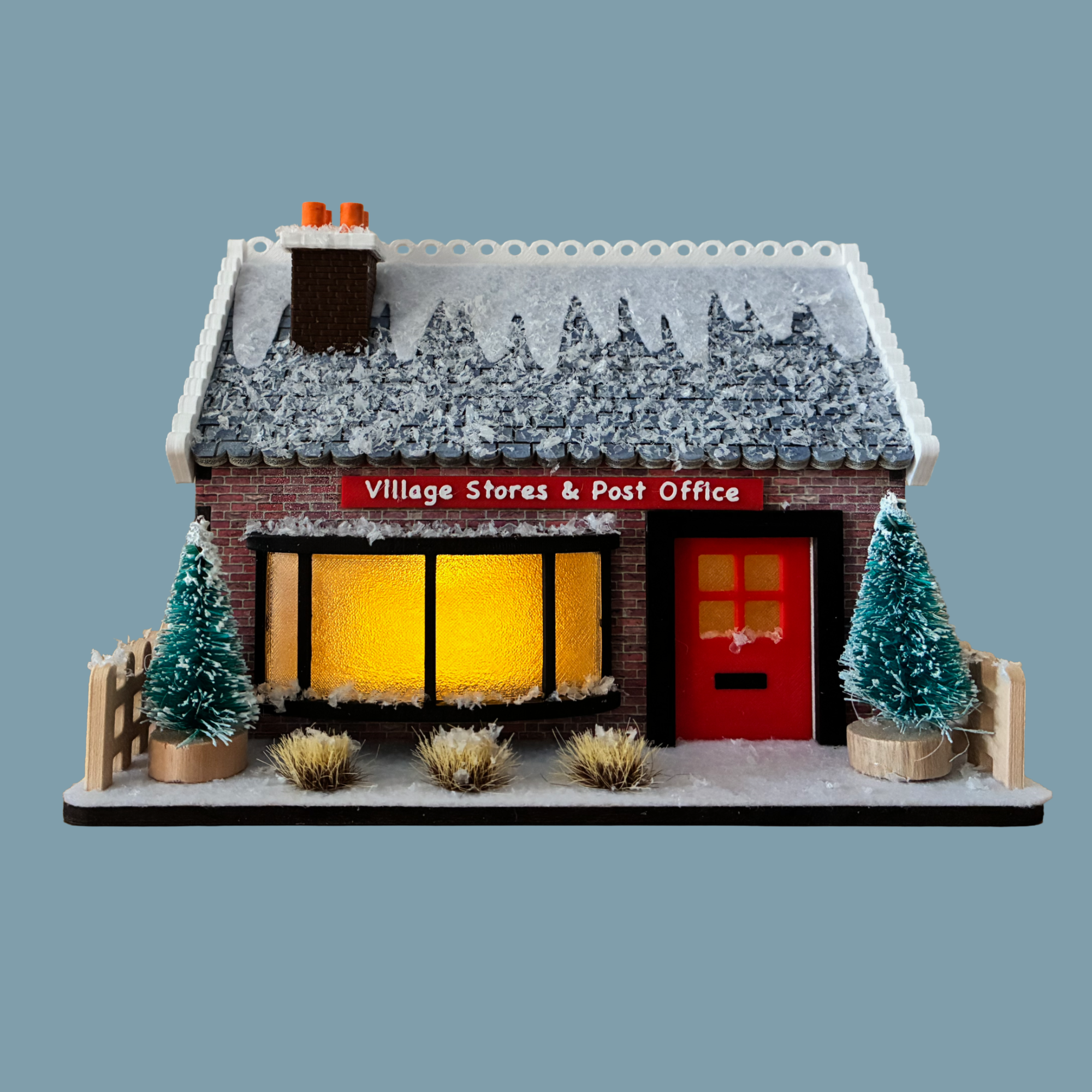 A miniature brick house model with a snow-covered gray roof, two glowing yellow windows, and a beige picket fence around it, shown on a plain blue-gray background.