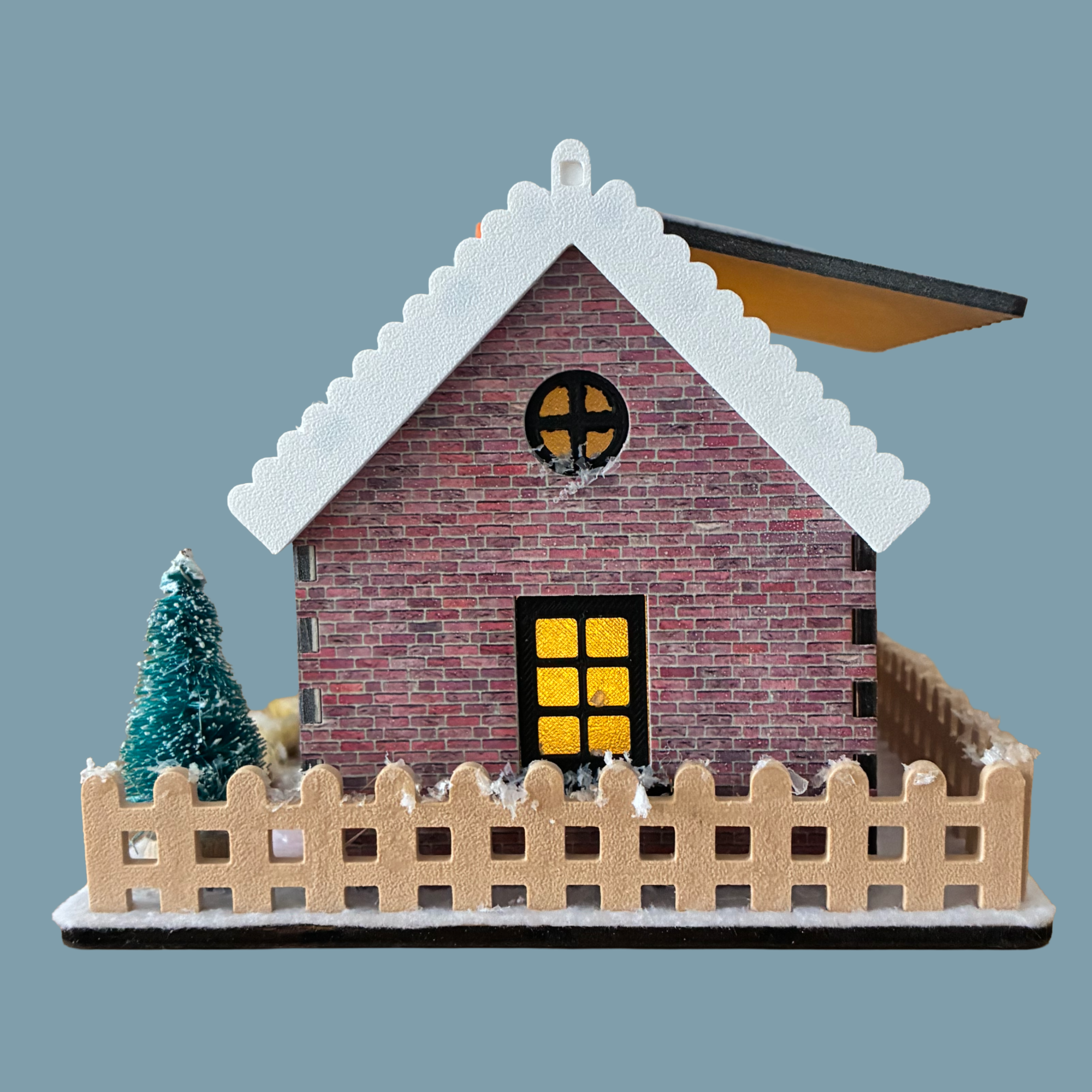 A miniature brick house model with a snow-covered gray roof, a glowing yellow window, and a beige picket fence around it, shown on a plain blue-gray background.